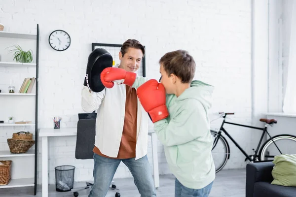 Smiling man in boxing pad training with son in gloves at home