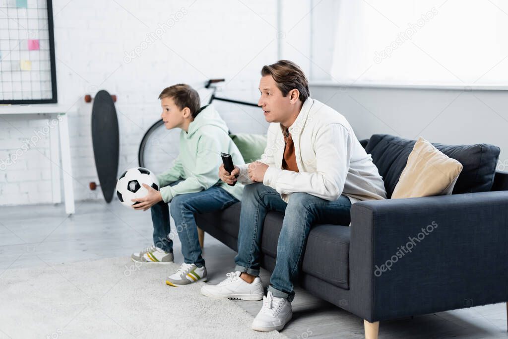 Focused man with remote controller watching football match near son at home 