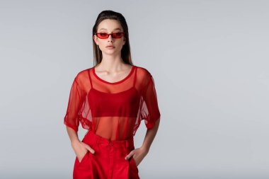 Kırmızı elbiseli ve güneş gözlüklü genç model. Gri cepli ellerle poz veriyor.