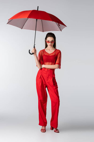 full length of young model in red trendy outfit and sunglasses standing under umbrella on grey