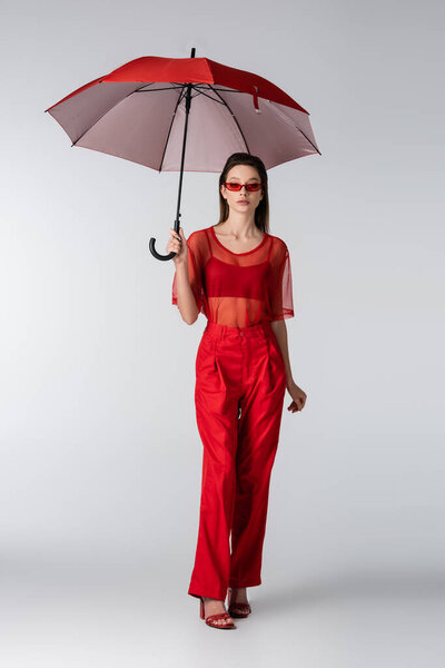 full length of young woman in red trendy outfit and sunglasses standing under umbrella on grey