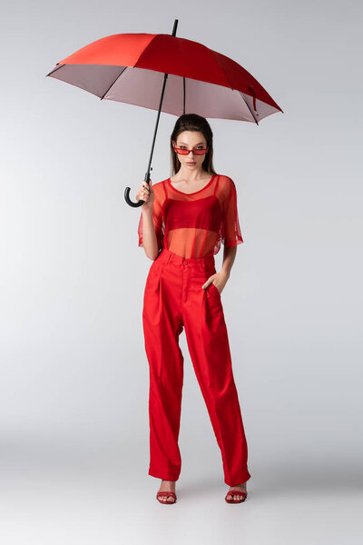 full length of young woman in red outfit and sunglasses standing with hand in pocket under umbrella on grey