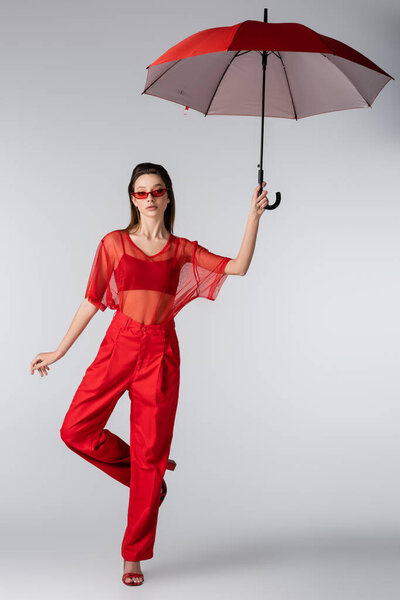 full length of young woman in trendy outfit and red sunglasses posing under umbrella on grey
