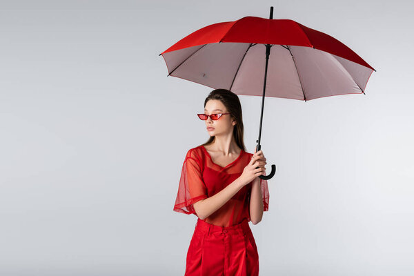 young model in trendy outfit and red sunglasses standing under umbrella isolated on grey
