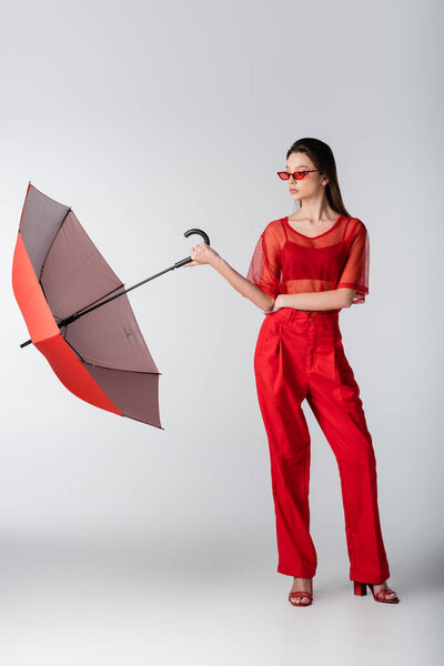 full length of young woman in trendy red outfit and sunglasses standing with umbrella on gray