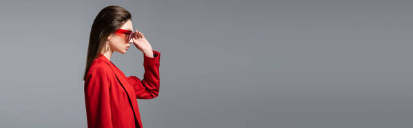 side view of young woman in trendy red suit adjusting sunglasses isolated on dark grey, banner