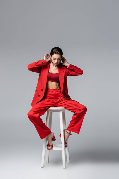 full length of brunette woman in red suit and shoes sitting on white chair on dark grey