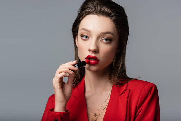 young woman in red blazer applying lipstick isolated on grey