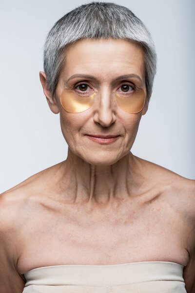middle aged woman in golden eye patches looking at camera isolated on grey