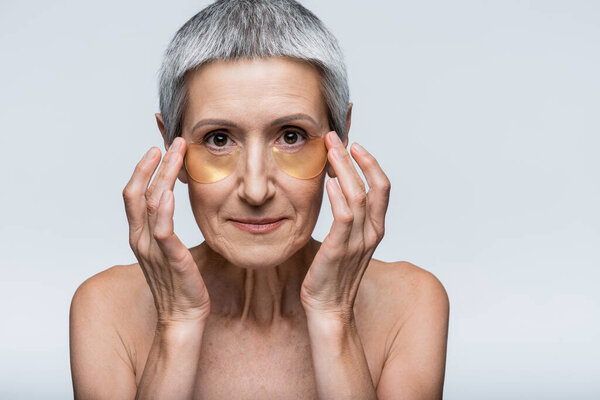 middle aged woman applying golden eye patches and looking at camera isolated on grey