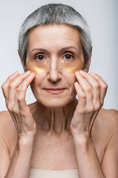 middle aged woman applying collagen eye patches and looking at camera isolated on grey