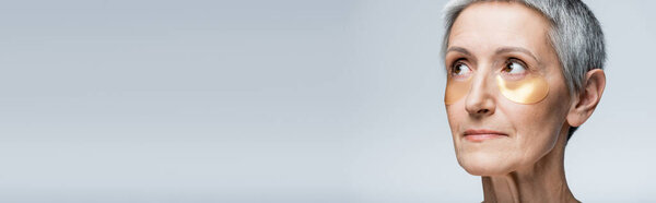 middle aged woman in golden eye patches looking away isolated on grey, banner