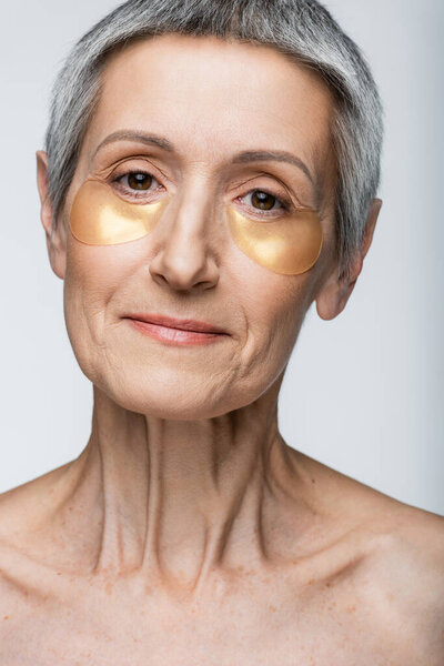 pleased middle aged woman in golden eye patches looking at camera isolated on grey