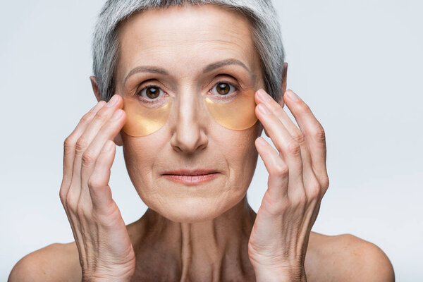 middle aged woman with bare shoulders applying golden eye patches isolated on grey