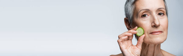 крупным планом женщины средних лет, держащей зеленый нарезанный огурец изолированный на сером, баннер