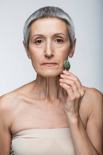 middle aged woman massaging face with jade roller isolated on grey 