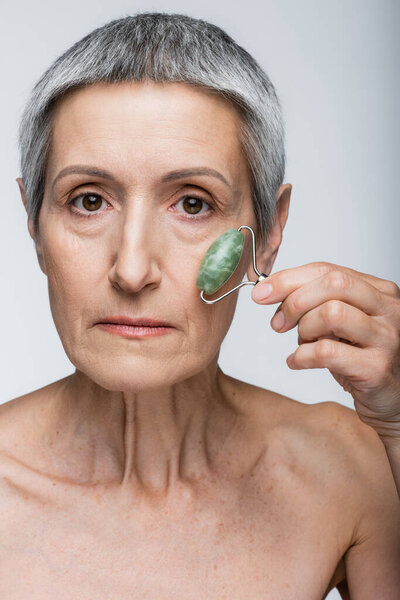 middle aged woman with grey hair massaging face with jade roller isolated on grey 