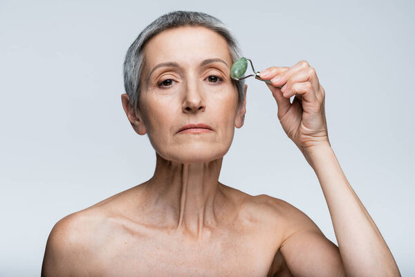 middle aged woman with bare shoulders massaging face with jade roller isolated on grey 