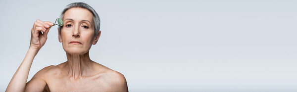 middle aged woman with bare shoulders massaging face with jade roller isolated on grey, banner