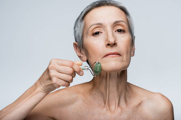 mature woman massaging face with jade roller isolated on grey 