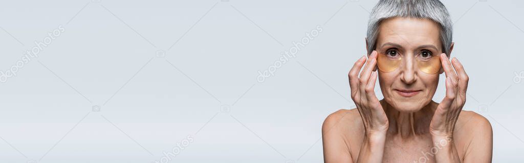 middle aged woman applying golden eye patches and looking at camera isolated on grey, banner
