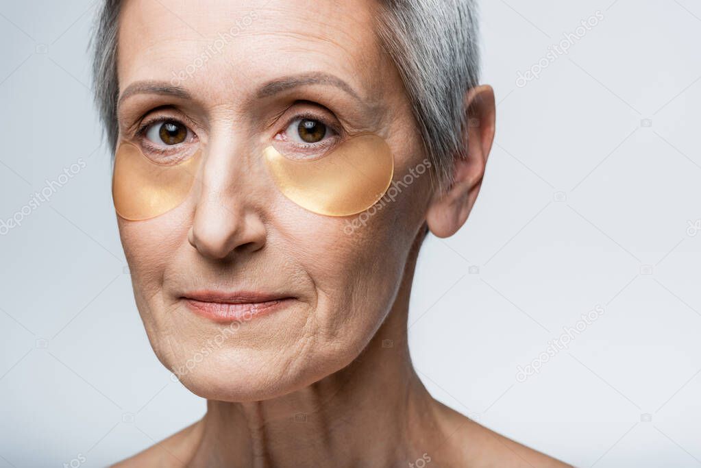 close up of middle aged woman in hydrogel eye patches looking at camera isolated on grey