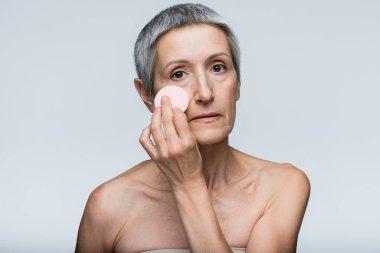 Gri üzerine izole güzellik süngeri ile makyaj vakfı uygulayan orta yaşlı kadın 