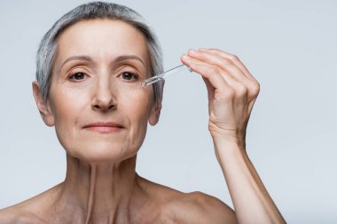 Gri saçlı orta yaşlı bir kadın elinde serumla gri renkli bir pipet tutuyor.