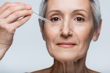 Gri saçlı orta yaşlı kadının, gri üzerine nemlendirici serum uygulamasını yakından izleyin.