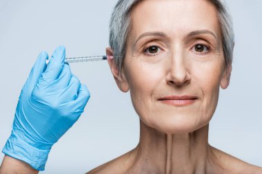 Plastik eldivenli güzellik uzmanı elinde güzellik enjeksiyonuyla gri üzerine izole edilmiş neşeli olgun bir kadının yanında.
