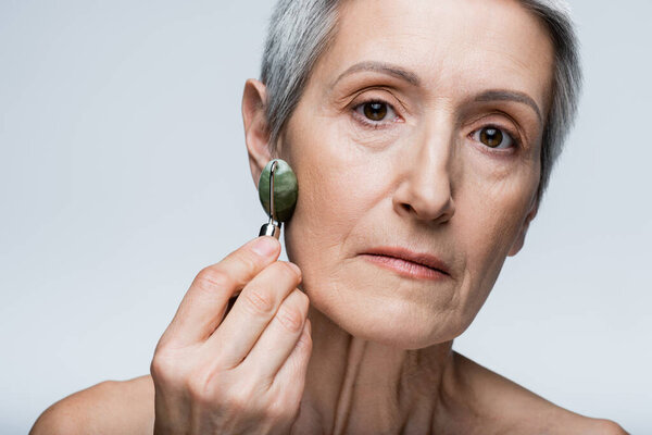 close up of mature woman massaging face with jade roller isolated on grey 