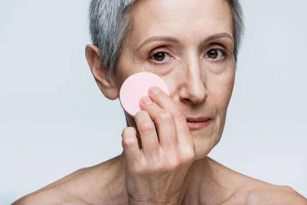 mature woman with bare shoulders holding beauty sponge and applying face foundation isolated on grey