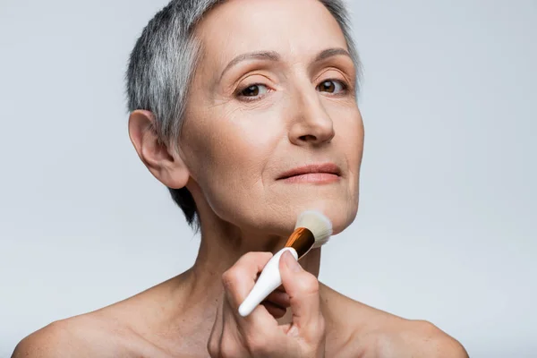 mature woman applying face powder with cosmetic brush isolated on grey