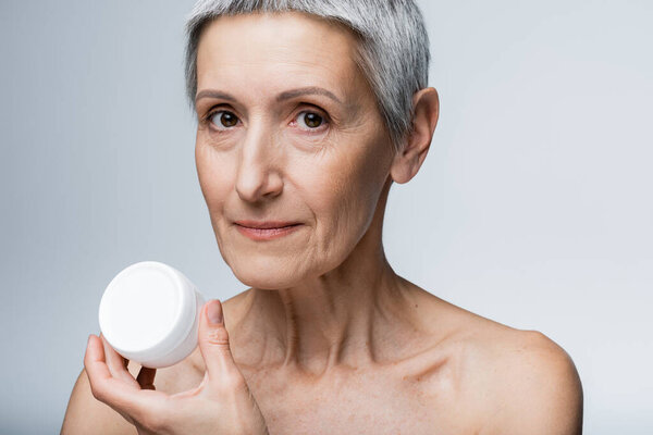 mature woman with grey hair holding container with face cream isolated on grey 