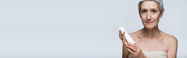женщина средних лет с седыми волосами с трубкой с лосьоном изолированы на сером, баннер