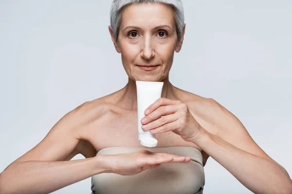 middle aged woman holding tube and applying lotion on hand isolated on grey