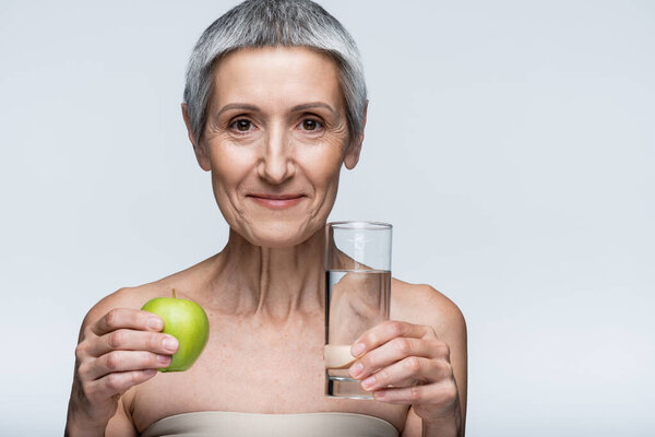 cheerful middle aged woman holding glass of water and green apple isolated on grey