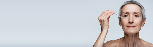 средневековая женщина с седыми волосами держит пипетку и применяет сыворотку, изолированную на сером, баннере