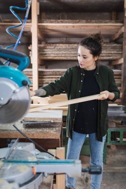 Woodworker holding plank near blurred miter saw on foreground in workshop  clipart