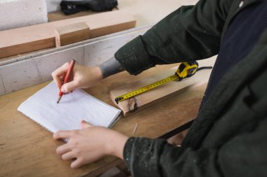 Cropped view of carpenter writing on notebook near wooden plank and tape measure in workshop  clipart