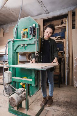 Young furniture designer holding board near band saw in workshop  clipart