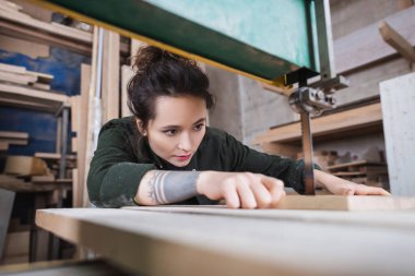 Brunette carpenter holding blurred wooden board and working in band saw in workshop  clipart