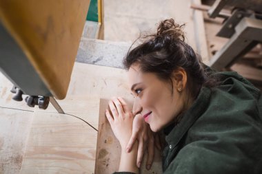 Side view of smiling furniture designer looking at plank and band saw in workshop  clipart