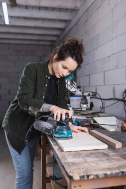 Brunette carpenter sanding wooden board near tools in workshop  clipart