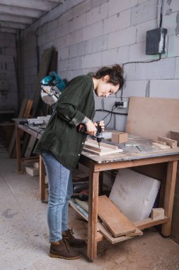 Side view of young furniture designer working with electric drill and wooden plank in workshop  clipart