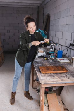 Tattooed carpenter taking photo on smartphone near wooden plank in workshop  clipart