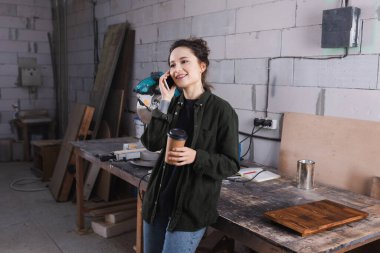 Happy woodworker holding paper cup and talking on cellphone near wooden board in workshop  clipart