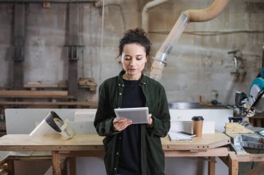 Genç marangoz atölyeye gitmek için güvenlik vizörü ve kahve yanında dijital tablet kullanıyor. 