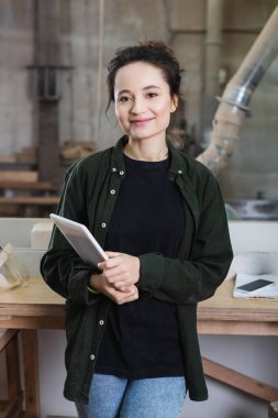 Pozitif marangoz dijital tablet tutuyor ve atölyedeki kameraya bakıyor 