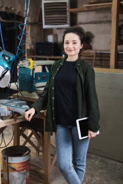 Positive carpenter holding digital tablet near miter saw in workshop  clipart